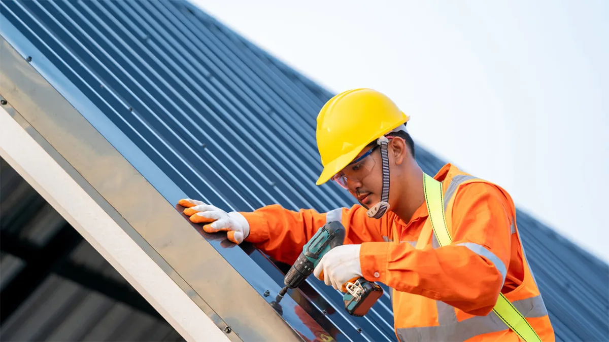 Construction worker drilling metal roof panel with safety equipment in place